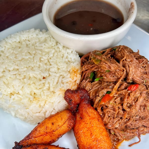 Latin-American dish with shredded beef, white rice, black beans, and fried plantains on a white plate.