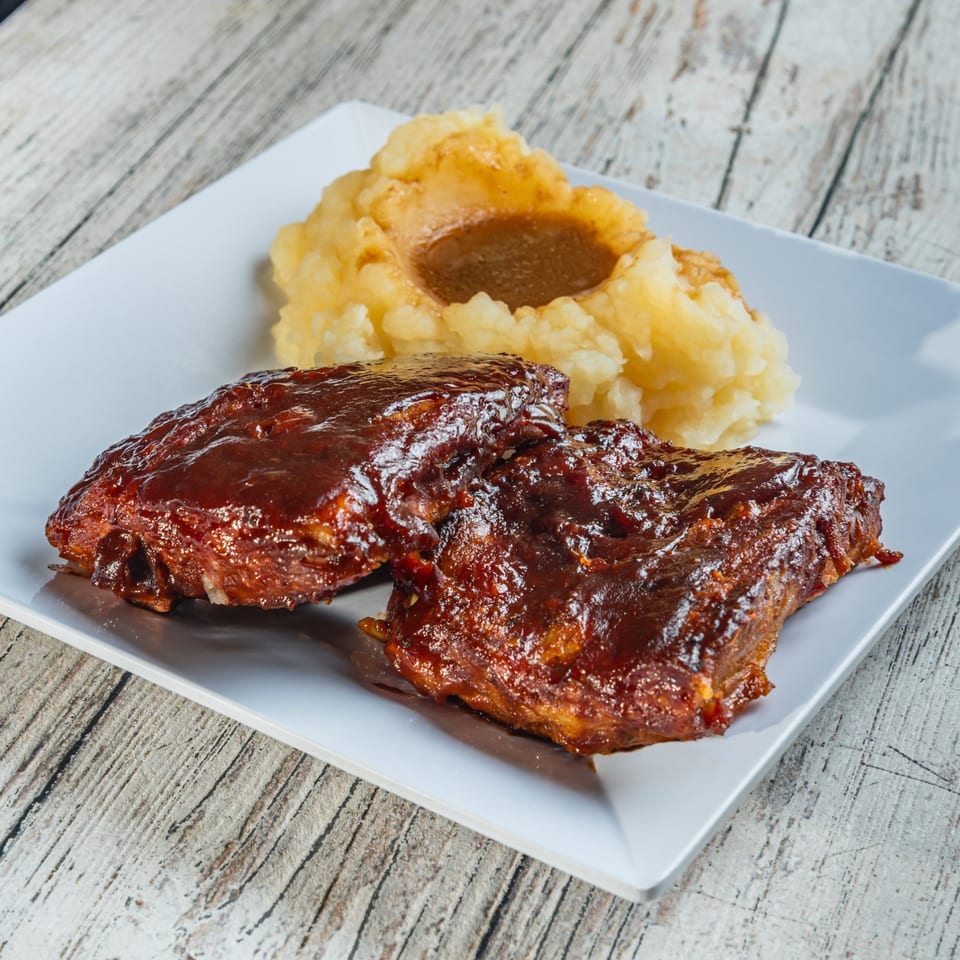 Two BBQ ribs with sauce and a side of mashed potatoes with gravy on a white plate.