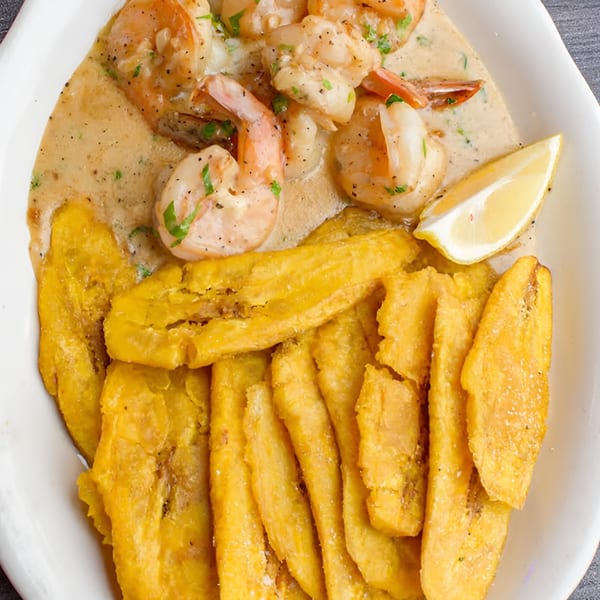 Shrimp in creamy sauce with fried plantains and a lemon wedge on a white plate.