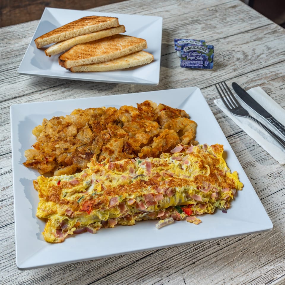 A breakfast plate with a ham and cheese omelet, hash browns, and toast with jelly.