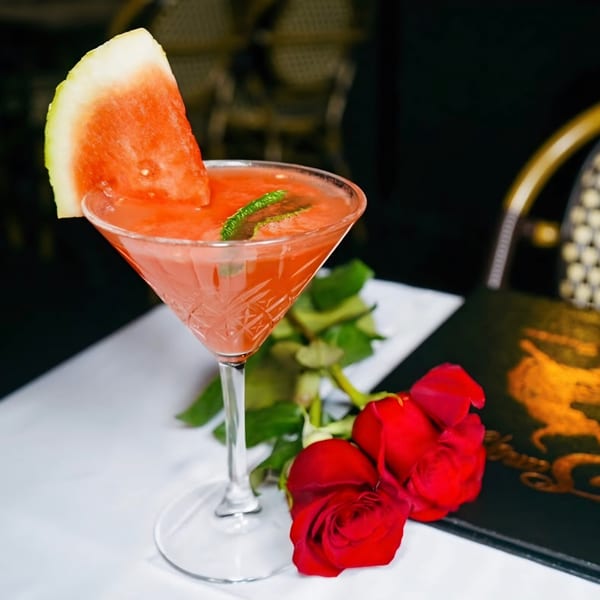 Watermelon cocktail in a martini glass with a watermelon slice, mint, and red roses on a table.