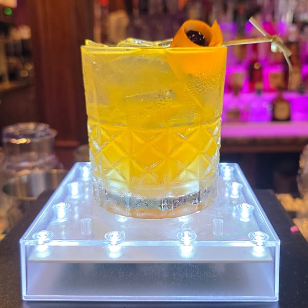 Yellow cocktail in a patterned glass with orange peel and cherry on a light-up coaster at a bar.