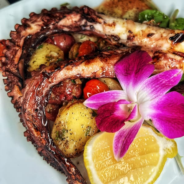 Grilled octopus with roasted potatoes, cherry tomatoes, lemon, and an orchid garnish on a white plate.
