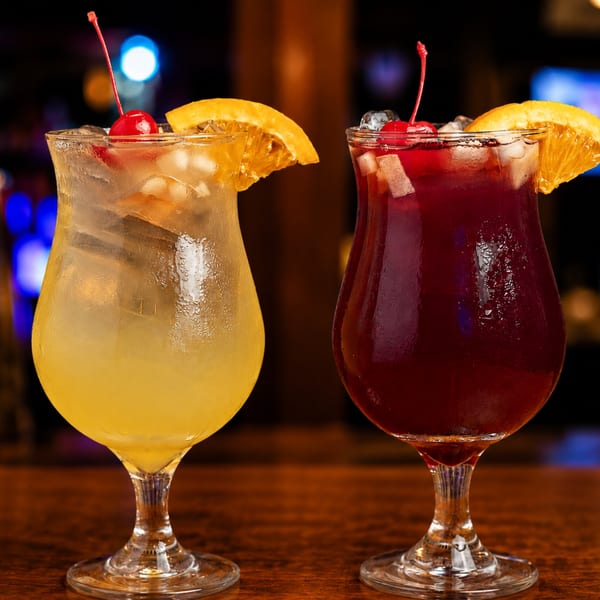 Two vibrant cocktails, yellow and red, with ice, orange slices, and cherries on a bar.