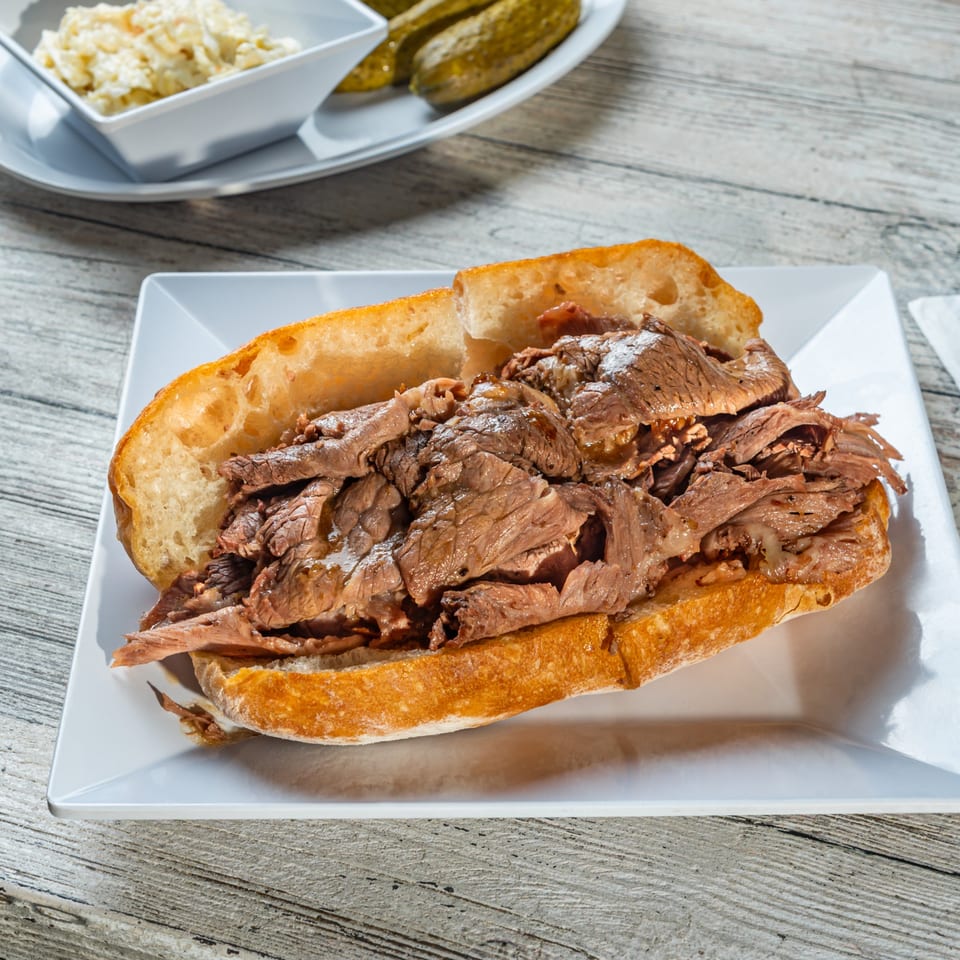 Hearty beef sandwich on a white plate with coleslaw and pickles in the background.
