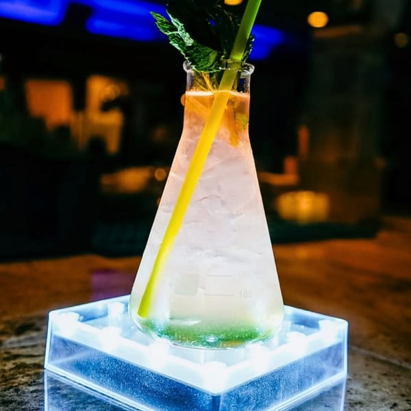 Cocktail in a science beaker with ice, mint, and a straw on a glowing blue coaster in a dark bar.