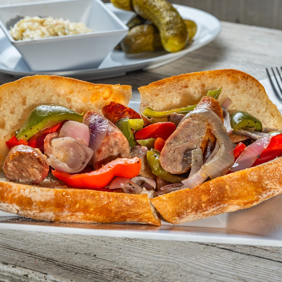 Sausage and pepper hero sandwich with onions, served with coleslaw and pickles.