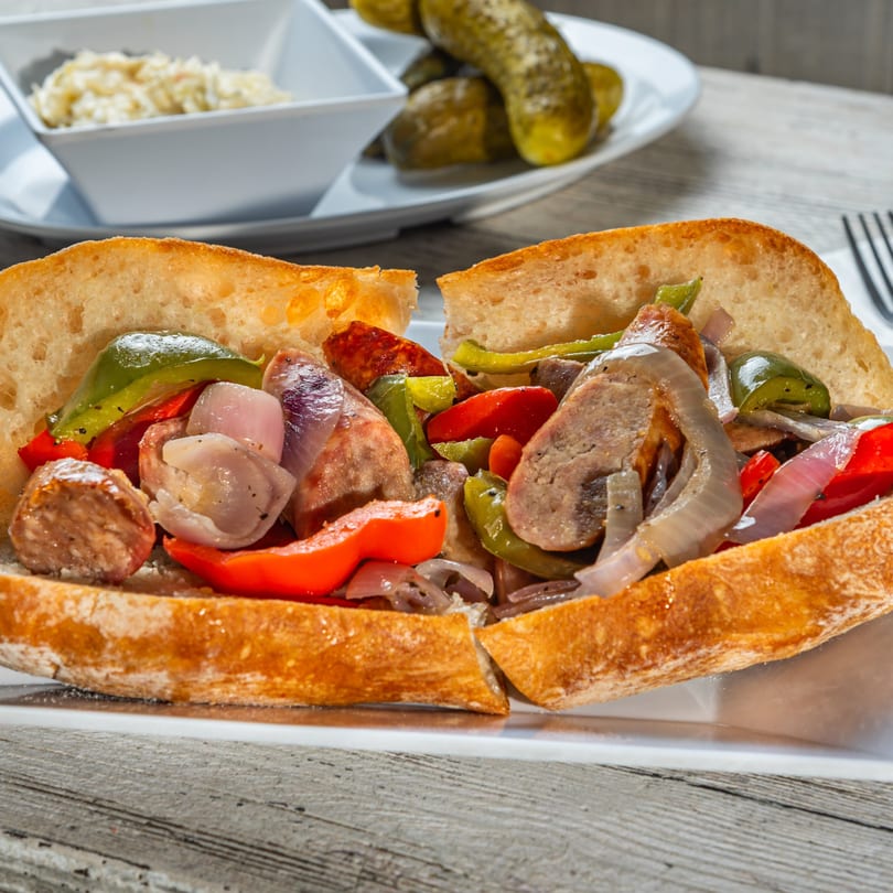 Sausage and pepper hero sandwich with onions, served with coleslaw and pickles.