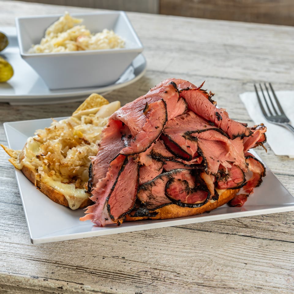Open-faced pastrami sandwich with melted cheese and sauerkraut, served with a side of coleslaw.