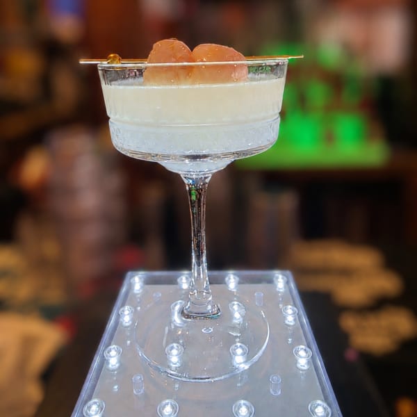 Elegant cocktail with lychee garnish in a coupe glass, displayed on a glowing stand in a bar setting.