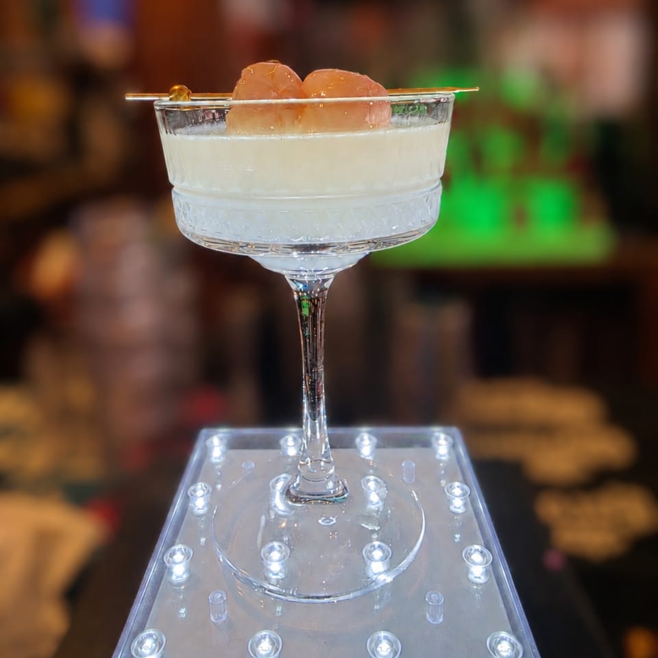 Elegant cocktail with lychee garnish in a coupe glass, displayed on a glowing stand in a bar setting.