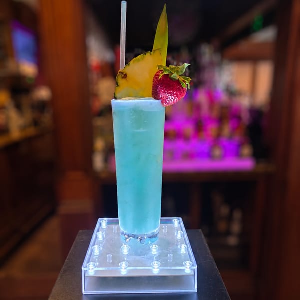 Tall blue cocktail garnished with pineapple and strawberry on a glowing coaster in a bar.