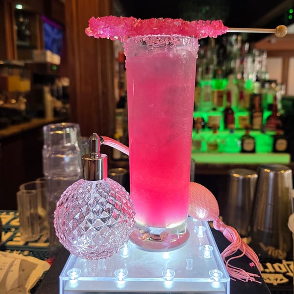 Tall pink cocktail with rock candy and a decorative bottle on a glowing stand at a bar.