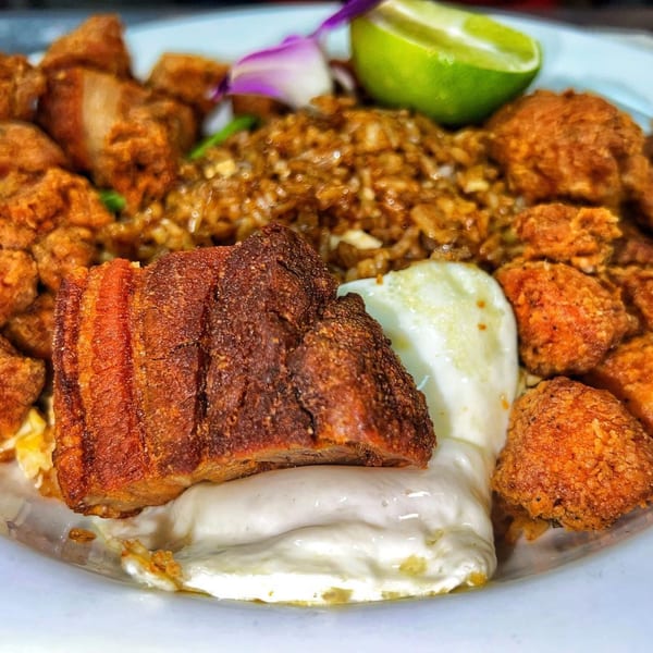 Crispy fried pork belly and chicken pieces with rice, a fried egg, and lime on a plate.