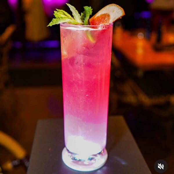 Vibrant pink cocktail with mint and grapefruit garnish, glowing from its base in a dark setting.